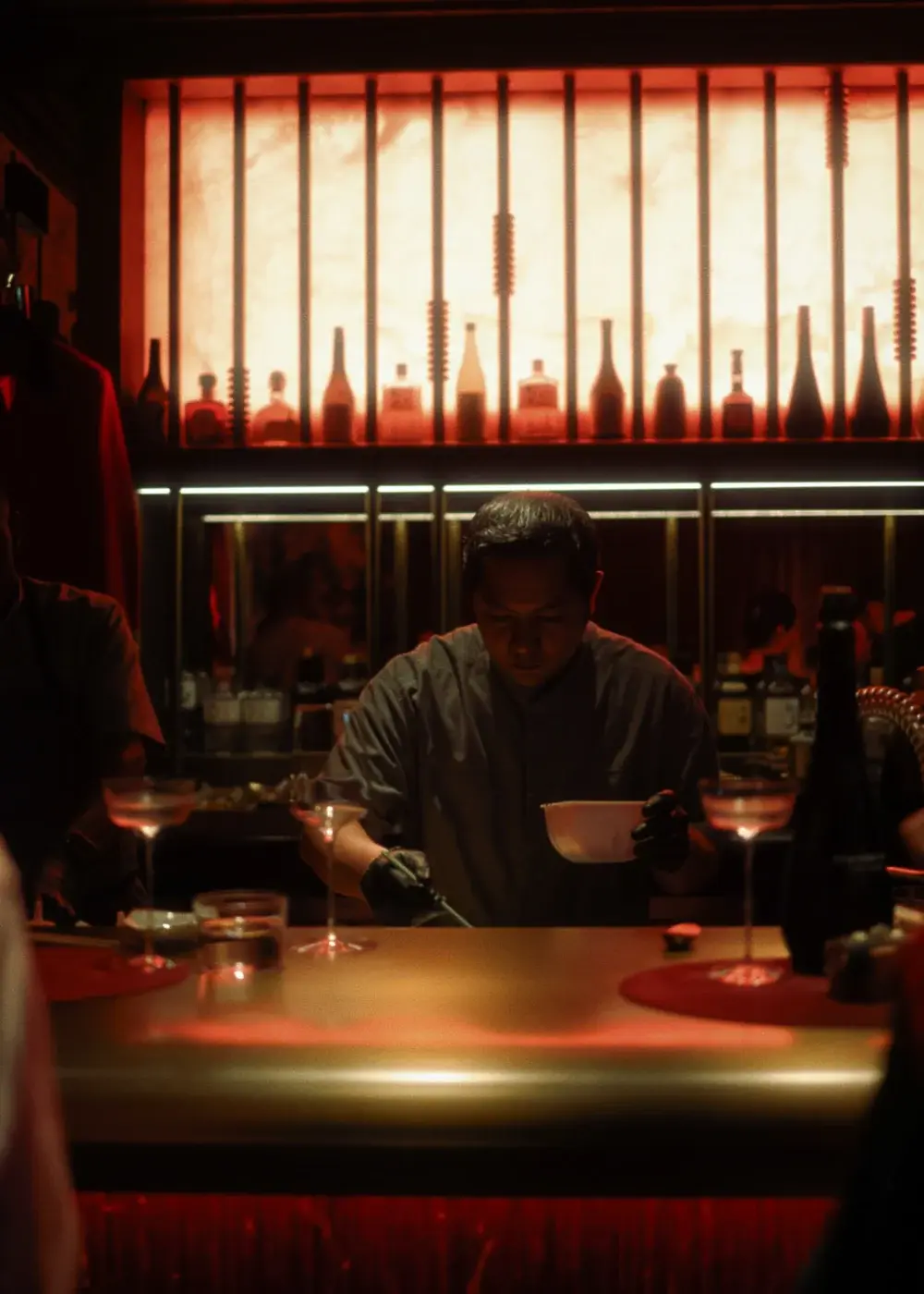 The chef plating at the counter at Himitsu, lit by the red sake wall behind him.