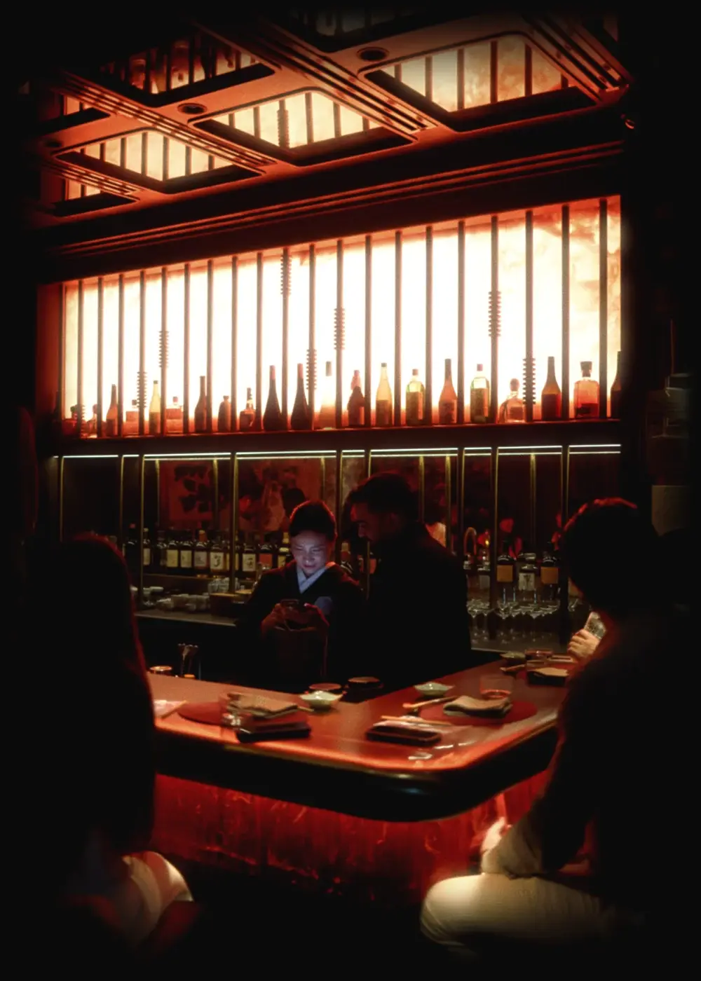 The Himitsu counter from across the room: a back-lit shelf of Japanese bottles above the bar, places set along the counter.